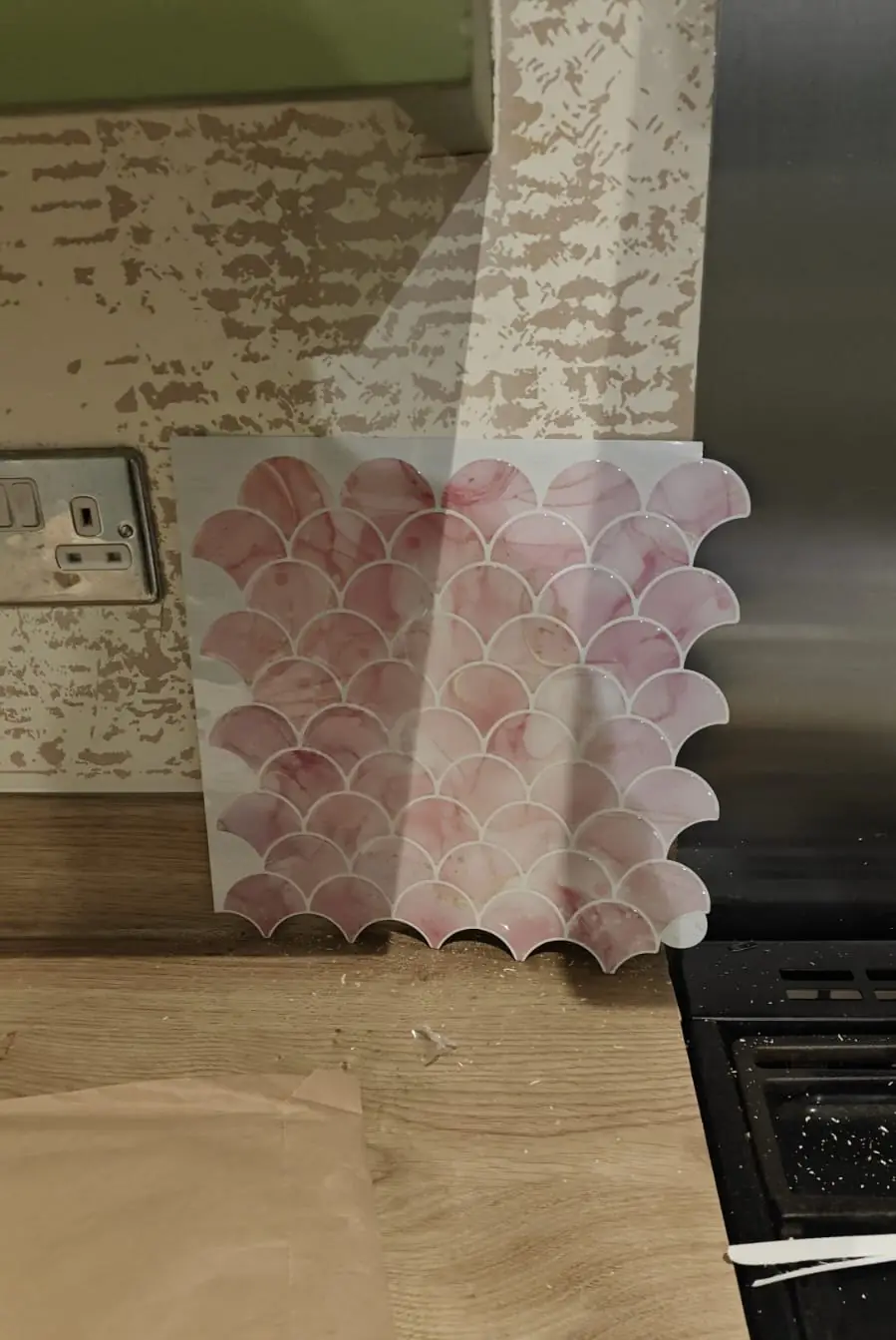 During: pink marble fish-scale peel-and-stick tile sample against the old wallpaper